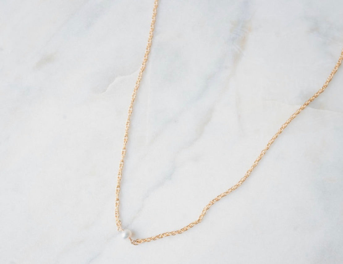 Gold chain necklace with a fresh water pearl in center on a light gray background